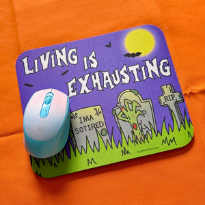 Mouse pad with Halloween graveyard design and text 'Living is Exhausting' on an orange surface.