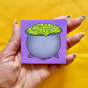 Hand holding a sticky notepad with a witch cauldron design against a yellow background